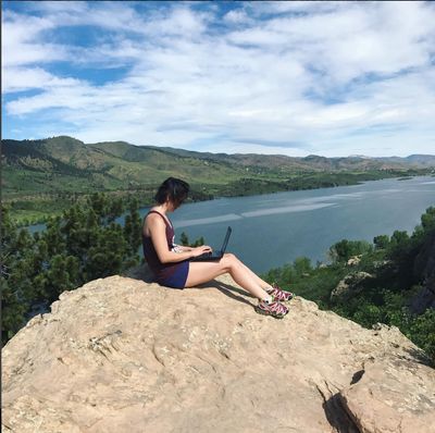 Brunette girl sitting on a rock working from a laptop. The background is a giant body of water and Horsetooth Rock formation set below a cloud filled blue sky.